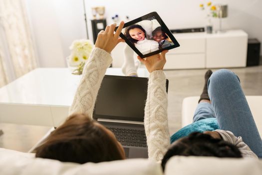 Two female friends making a selfie with a digital tablet sitting on the couch at home. Multiethnic women.