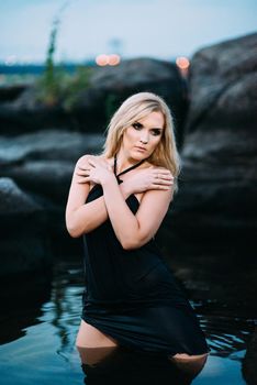 blonde girl in a black dress with blue eyes on the stone coast of the river