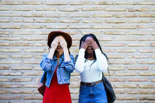 Two friends covering their eyes in an urban background.. Multiethnic women.