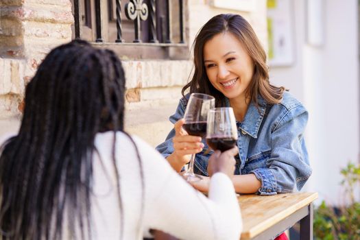 Two friends making a toast with red wine sitting at a table outside a bar. Multiethnic women.
