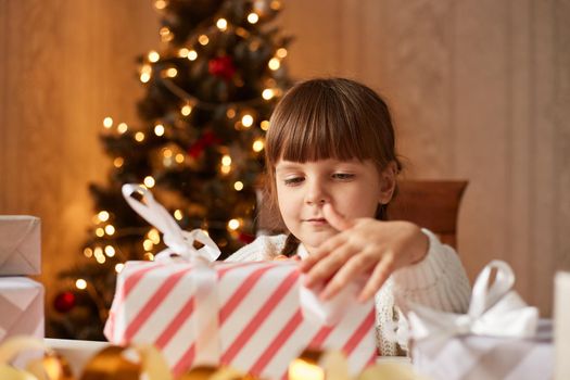 Portrait of charming adorable little girl packing present boxes for Christmas, dark haired female child with curious expression preparing for winter holidays.