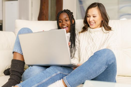 Two female student friends sitting on the couch at home using a laptop. Multiethnic women.