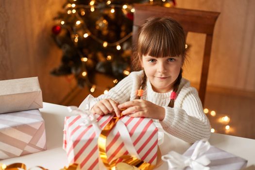 Indoor shot of charming positive adorable female child wearing white sweater looking at camera while packing Christmas presents, celebrating New year eve.