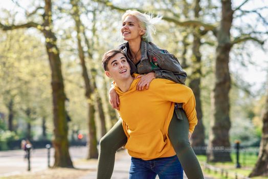 Funny couple in a urban park. Boyfriend carrying his girlfriend on piggyback. Love and tenderness, dating, romance. Lifestyle concept