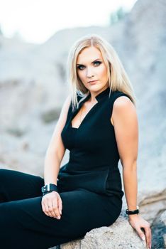 blonde girl in a black trouser suit with blue eyes on the background of a granite quarry from gray rubble