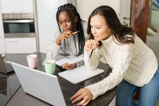 Two college girls studying together at home with laptops while drinking coffee. Multiethnic women.