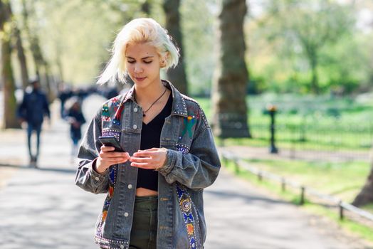 Young urban woman with modern hairstyle using smartphone walking in street in an urban park in London, UK.