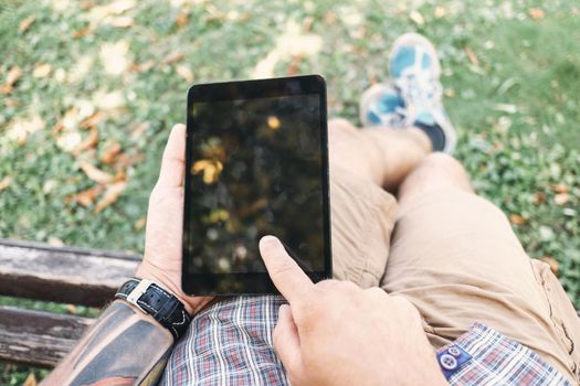 Young man sitting in park and using digital tablet, point of view.