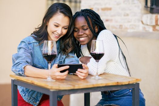 Two women looking at their smartphone together while having a glass of wine on the terrace of a bar. Multiethnic women.