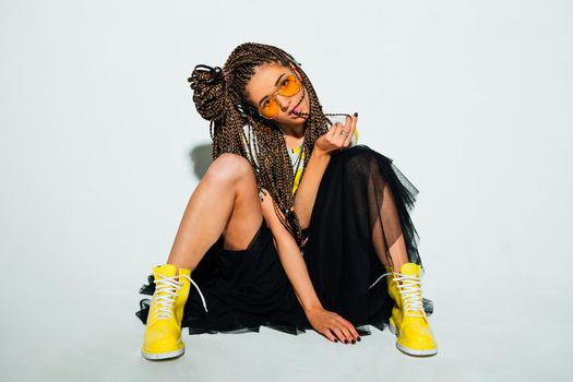 Portrait of a young stylish woman with braided hair dressed in white t-shirt, black skirt, yellow boots and sunglasses posing on the white background.