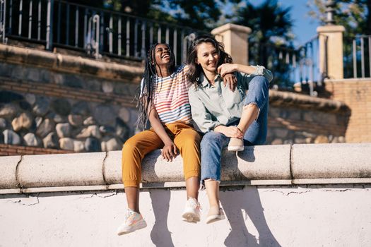 Two friends having fun together on the street sitting on a urban wall. Multiethnic women.