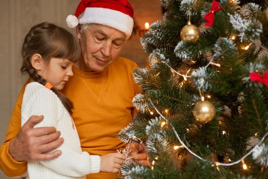 Family celebrating Christmas at home, grandad and grandchild decorating christmas tree together, look concentrated, wearing casual clothing, mature man in santa claus hat.