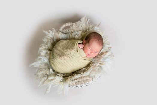 Cute newborn baby wrapped in beige blanket smiles while sleeping on the soft basket, top view.