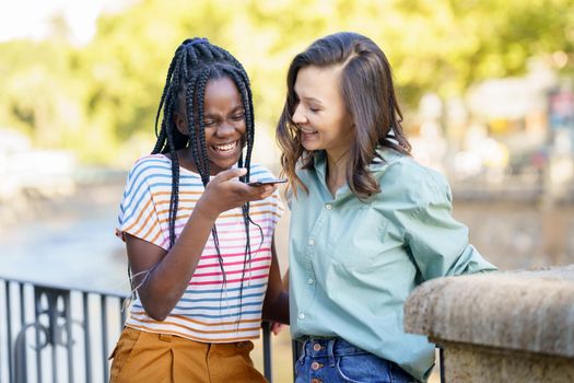 Two female friends recording a voice note with smartphone outdoors. Multiethnic friends.