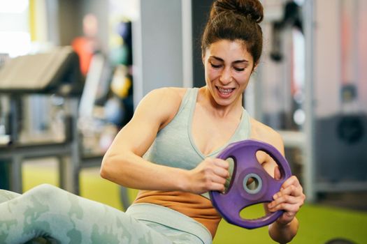 Sporty young female, wearing sportswear, on yoga mat doing situps with plate weights in gym. Fitness woman doing abs crunches.