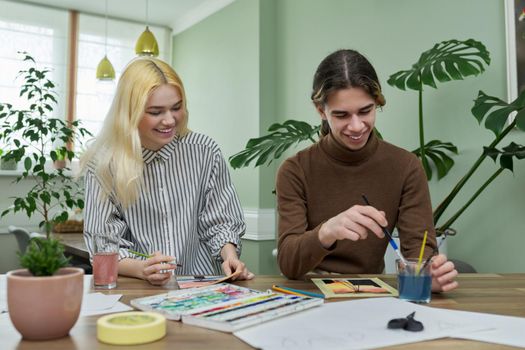 Teenagers painting with watercolors. Couple of guy and girl sitting at table with paints, brushes, drawings. Happy teens talking laughing creative. Creativity, hobby, leisure concept