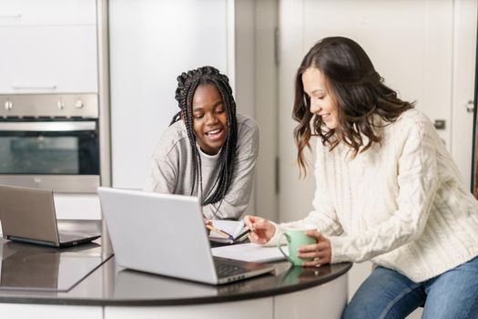 Two college girls studying together at home with laptops while drinking coffee. Multiethnic women.