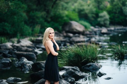 blonde girl in a black dress with blue eyes on the stone coast of the river