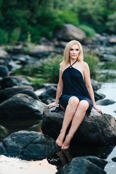 blonde girl in a black dress with blue eyes on the stone coast of the river