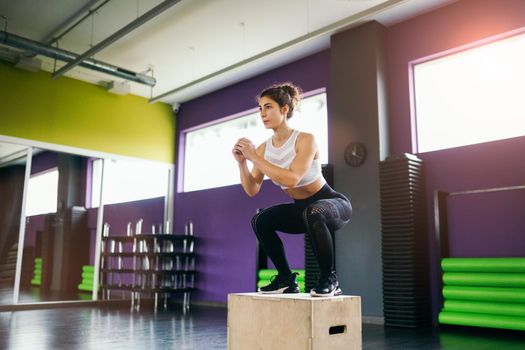 Athletic woman doing squats on box as part of exercise routine. Caucasian female doing box jump workout at gym.