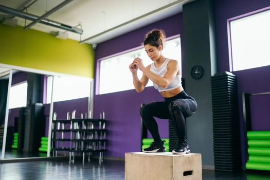 Athletic woman doing squats on box as part of exercise routine. Caucasian female doing box jump workout at gym.