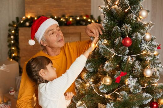 Family celebrating Christmas, grandpa and child girl with pigtails decorating Christmas tree, female king in white sweater and senior man in orange jumper posing indoor in festive room.
