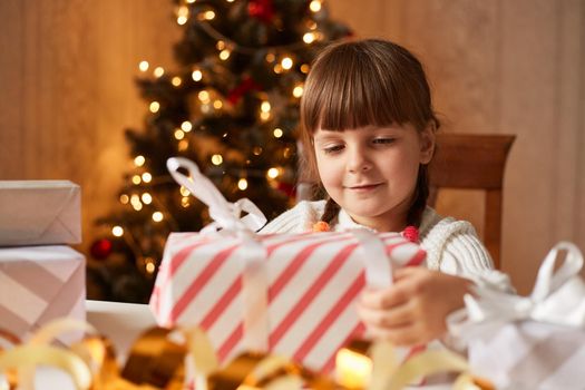 Smiling little girl with dark hair and pigtails packing Christmas presents for her friends, sitting at table, having festive mood, expressing positive emotions.