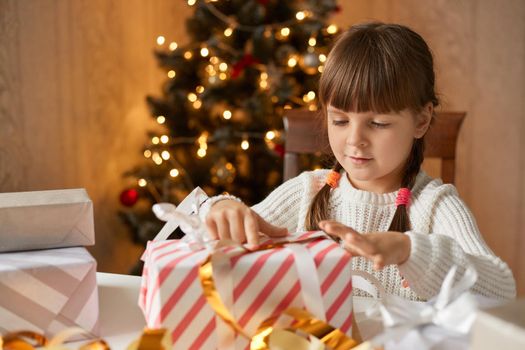 Happy girl opening gift box while sitting at table, female kid with pigtails looking at striped box with interest, little girl posing in room with x,mas tree on background.