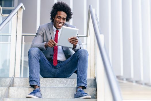 Smiling Black Businessman using a digital tablet sitting near an office building. Man with afro hair.