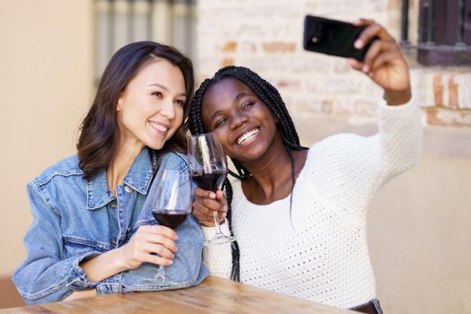 Two women making a selfie with a smartphone while having a glass of wine on the terrace of a bar. Multiethnic women.