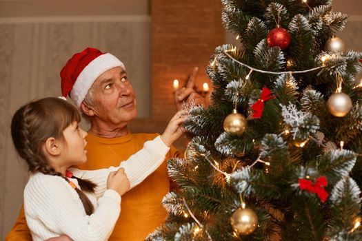 Grandfather and granddaughter decorating christmas tree, family wearing casual style clothing looking at beautiful z mas tree, celebrating new year eve together.