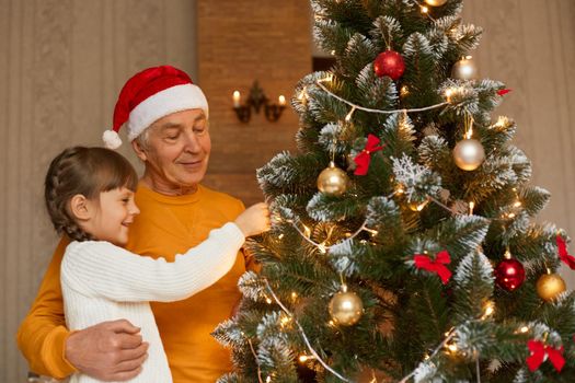 Grandfather and granddaughter decorating xmas tree, looking at fir tree with smile, child in white jumper, senior man wears yellow shirt and santa claus hat, posing indoor in festive room.