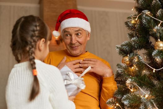 Senior man wearing orange shirt and santa claus hat, getting christmas present from his granddaughter, being astonished to get present from grandchild, posing indoor near fir tree.