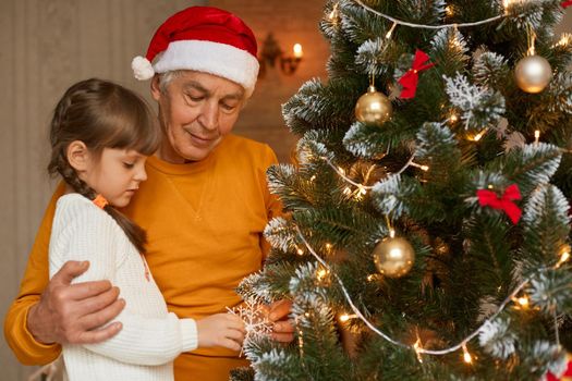 Happy family posing indoor near xmas tree, grandfather in orange shirt and santa claus hat hugging his charming granddaughter decorating fir tree, celebrating New year.