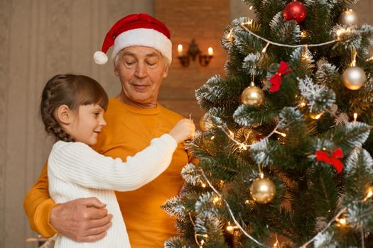 Family decorating christmas tree at home, cute little female child wearing white sweater decorate xmas tree with her grandfather, posing indoor, celebrating new year eve.