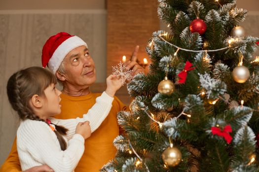 Smiling grandad with little granddaughter decorating christmas tree at home, wearing casual clothing, posing indoor, looking at christmas tree, New Year eve.