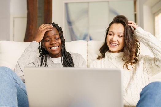 Two female student friends sitting on the couch at home using a laptop. Multiethnic women.