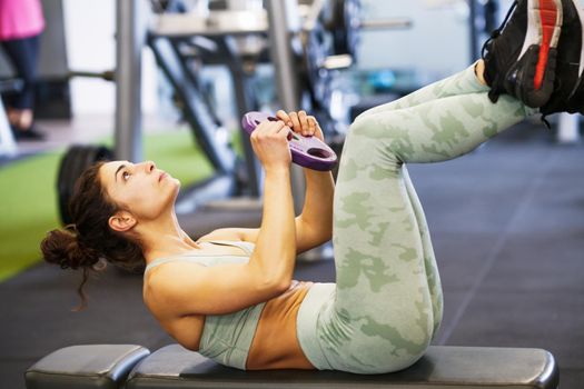 Sporty young female, wearing sportswear, on yoga mat doing situps with plate weights in gym. Fitness woman doing abs crunches.