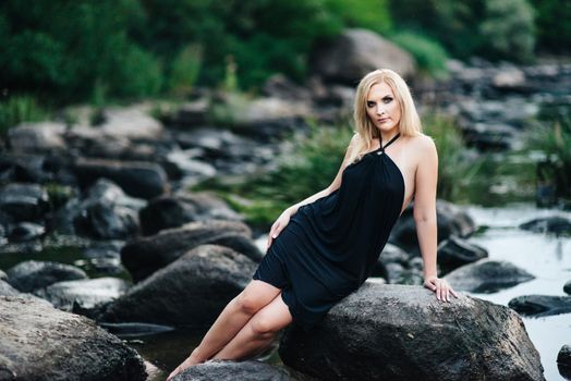 blonde girl in a black dress with blue eyes on the stone coast of the river