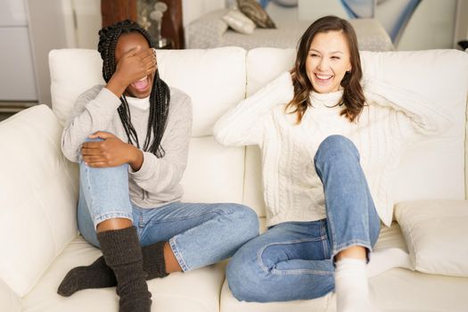 Two female student friends laughing together on the couch at home. Multiethnic women.