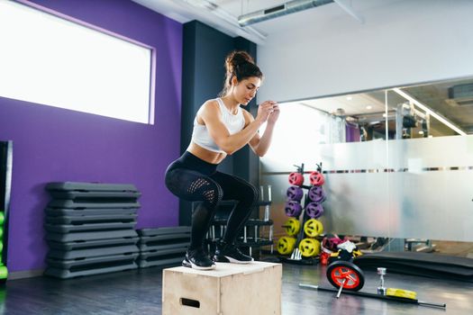 Athletic woman doing squats on box as part of exercise routine. Caucasian female doing box jump workout at gym.