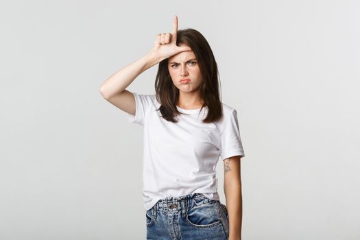 Sad and disappointed brunette girl showing loser gesture and sulking, white background.