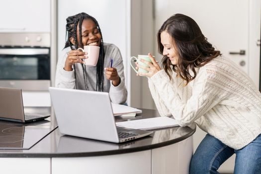 Two college girls studying together at home with laptops while drinking coffee. Multiethnic women.