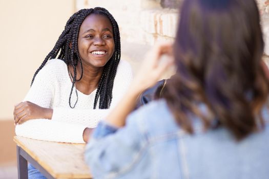 Two friends talking sitting at a table outside a bar. Multiethnic women.