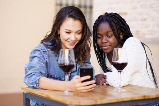 Two women looking at their smartphone together while having a glass of wine on the terrace of a bar. Multiethnic women.