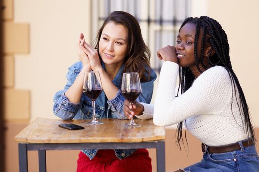 Two friends drinking red wine sitting at a table outside a bar. Multiethnic women.