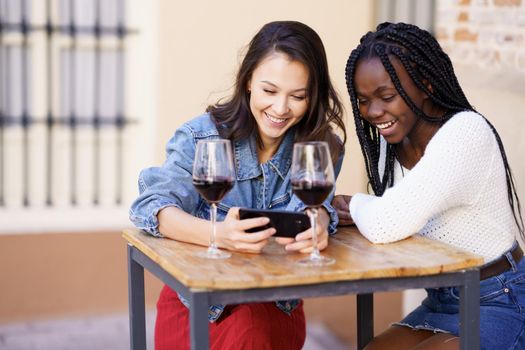 Two women looking at their smartphone together while having a glass of wine on the terrace of a bar. Multiethnic women.