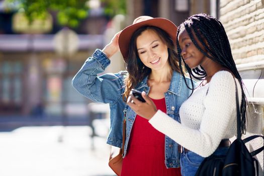 Two friends looking at their smartphone together on the street. Multiethnic women.
