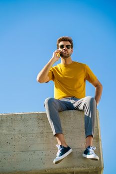 Young man using his smartphone sitting on a ledge outside. Guy wearing sunglasses.