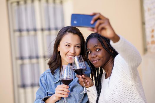 Two women making a selfie with a smartphone while having a glass of wine on the terrace of a bar. Multiethnic women.